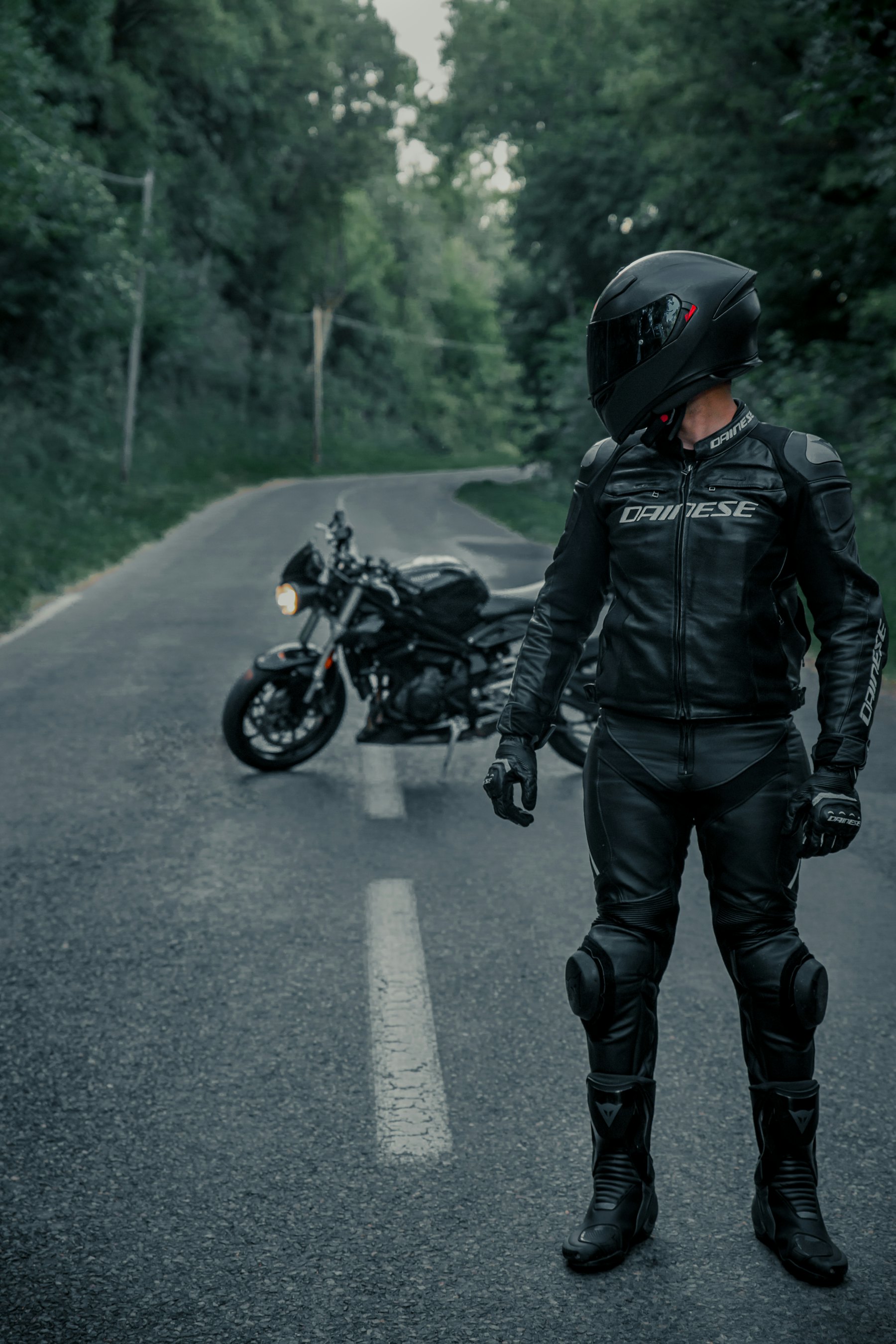 A rider in full black leather motorcycle gear standing beside a motorcycle on a quiet forest road, shot in high-contrast black and white