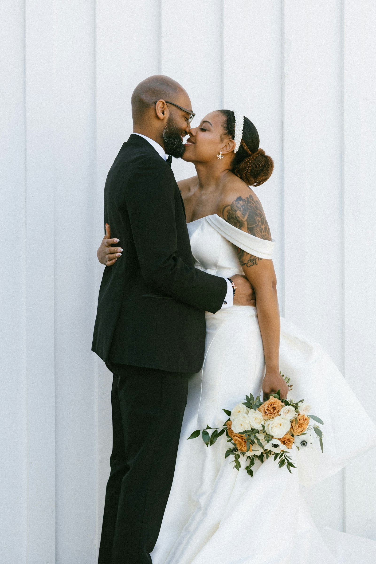 A groom in a black suit kissing his tattooed bride against a plain wall, shot in stark black and white
